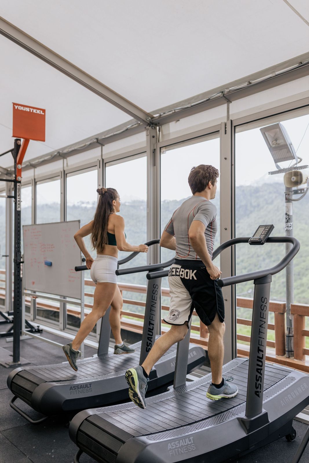 Two athletes running on curved manual treadmills beside large windows with a mountain view