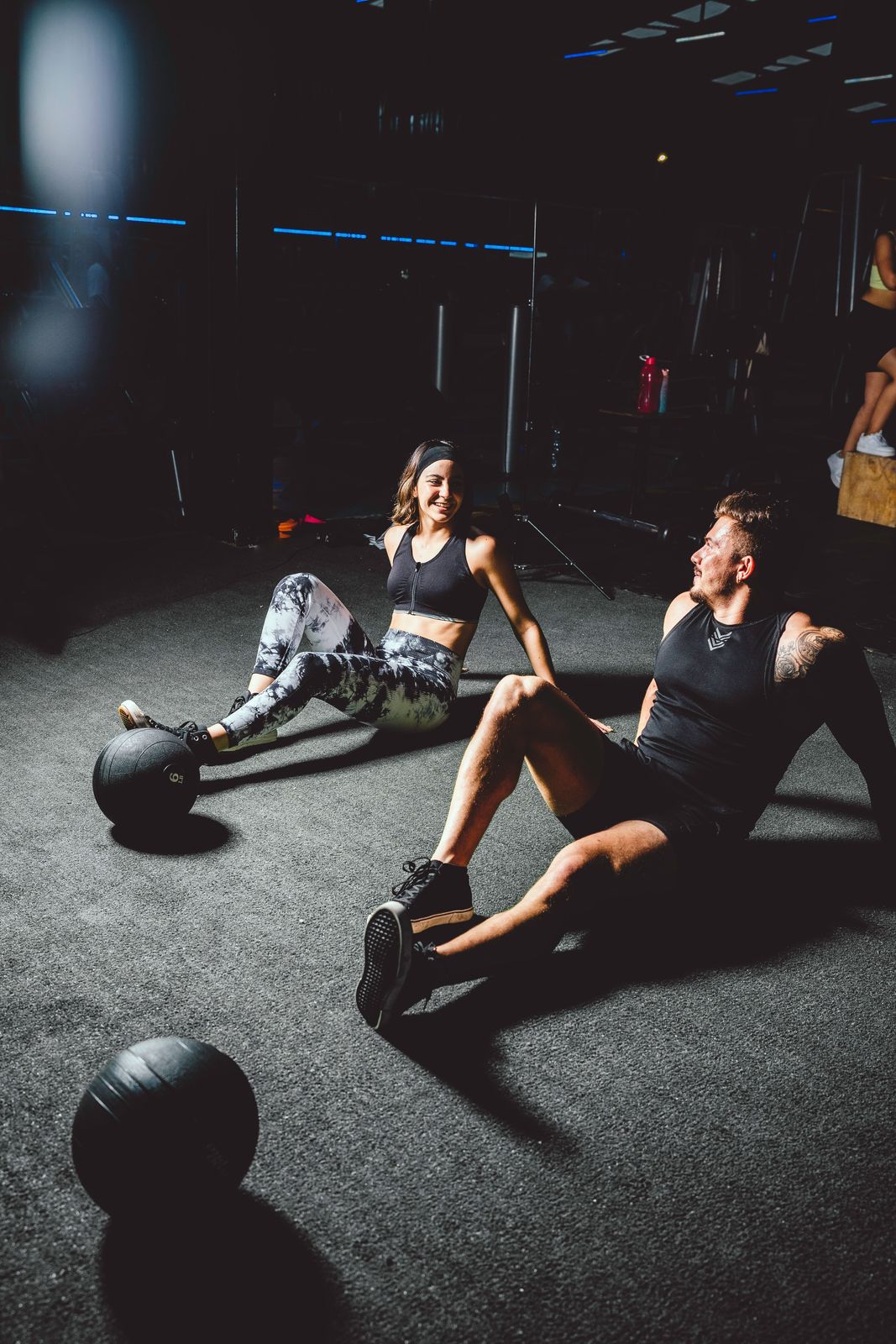 Two training partners sitting on a gym floor between medicine balls, recovering and talking after a workout